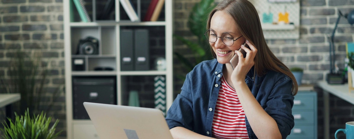 A woman smiles as she talks on the phone and looks at her laptop.